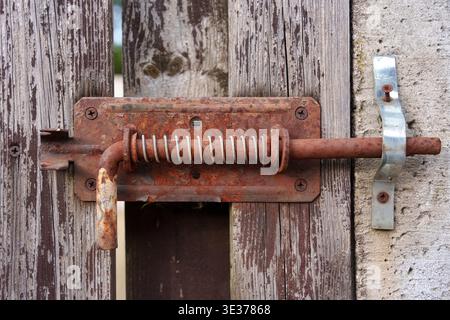Close-up of an old, rusty metal sliding bolt latch with a spring mechanism mounted on a weathered wooden fence door Stock Photo
