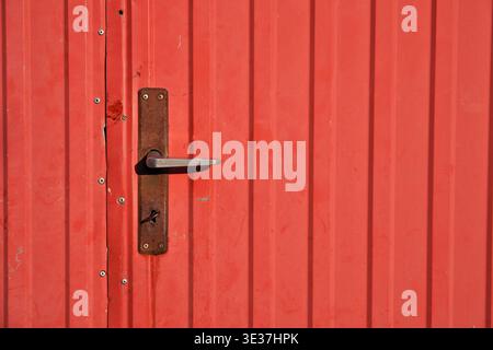 Close-up of a rustic, rusty metal door handle with a keyhole mounted on a bright red corrugated metal door Stock Photo