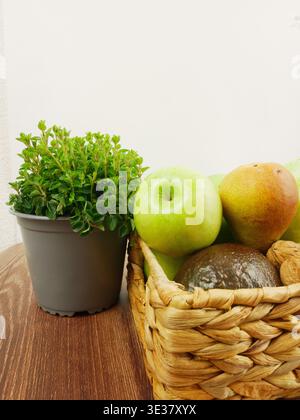 Green apples and pears are arranged in a basket next to a potted herb Stock Photo