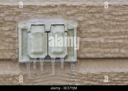 Close-up of protected electrical outlets mounted on outside wall of home and covered with thick ice and hanging icicles after freezing rain in late wi Stock Photo