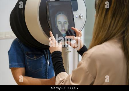 Aesthetic center technician using a modern skin analysis device with a tablet display, performing a facial mapping on a woman to provide personalized Stock Photo