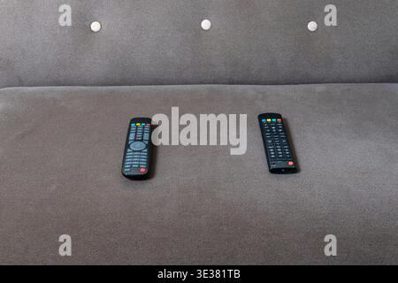 Two black remote controls rest on a textured grey sofa. for home entertainment, leisure, relaxation, technology, and modern living. Stock Photo