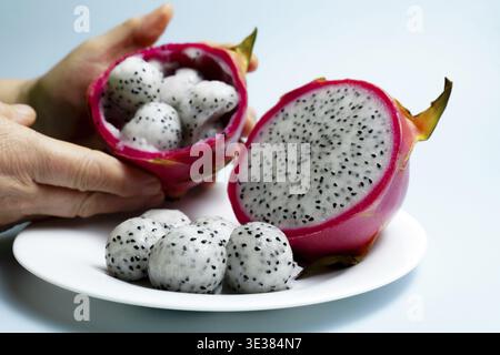 Hands preparing fresh pitaya, removing the seed filled white pulp using a spoon and placing it on a white plate, with another sliced dragon fruit on a Stock Photo