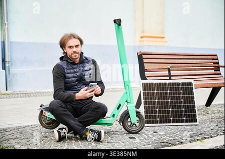 Portrait of male person sits on electric scooter with solar panel and smartphone while charging. Integration of renewable energy in personal transportation, highlighting sustainable urban lifestyle. Stock Photo