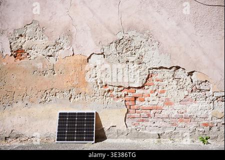Photovoltaic solar panel at weathered, textured wall with peeling paint. Juxtaposition of modern renewable energy technology against rustic, decaying backdrop highlights contrast between old and new. Stock Photo