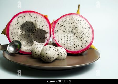 Dragon fruit halves displayed with scoops of its white flesh and tiny black seeds on a plate, highlighting its vibrant pink peel and healthy tropical Stock Photo