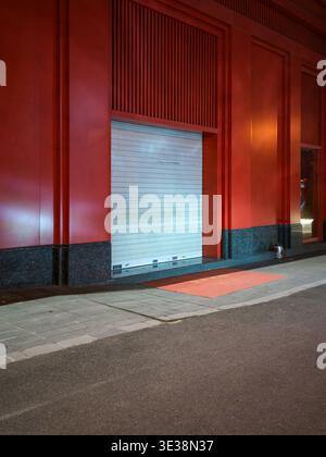 A closed metal roller shutter door set into a striking red building facade along an empty asphalt city street at night. The scene represents commercia Stock Photo