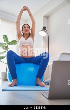 Pregnant woman balancing on a fitness ball and stretching arms during a home workout class Stock Photo