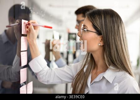 Two young colleagues writing on sticky notes attached on board Stock Photo