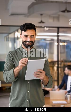 Smiling Middle Eastern businessman using digital tablet stands in office Stock Photo
