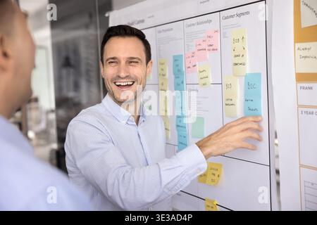Laughing businessman showing male colleague sticky note on agile board Stock Photo
