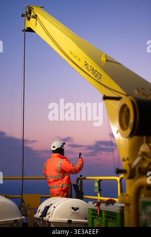 Offshore worker using a smartphone on a platform deck at sunset, with offshore wind turbines in the distance. Stock Photo