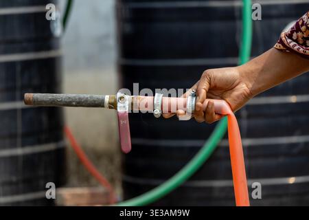 Close-up of a human hand holding a metal water pipe with a red handle ball valve and orange plastic hose outdoors. Perfect for plumbing services, wate Stock Photo
