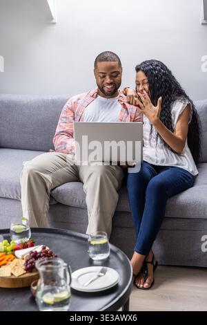 Couple sitting on gray sofa sharing silver laptop on man's lap, woman showing ring Stock Photo