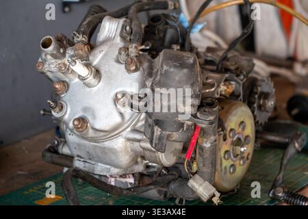 Old motorcycle engine on workbench, parts exposed, tools nearby, metal shiny, wires attached, gear visible, green cutting mat under Stock Photo