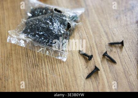 Close up of small black screws and plastic bag of hardware fasteners on wooden table. Metal equipment for construction, carpentry, and DIY home improv Stock Photo