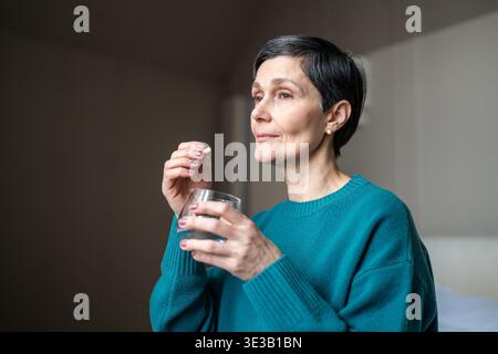 Senior woman taking special medicine to cure illness, holding glass of water and pill supplement. Stock Photo