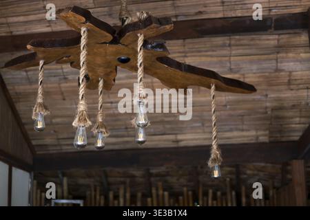 Unique rustic wooden chandelier with hanging light and rope detail. This cozy handmade lamp creates warm atmosphere and decorates natural bamboo inter Stock Photo