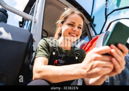 Happy female farmer using her smartphone near a tractor outdoors. Stock Photo