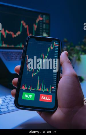 Close-up of a trader hand holds a smartphone with trading app, financial charts or cryptocurrency prices on screen, showing buy and sell buttons during online stock buying or crypto investing. Stock Photo