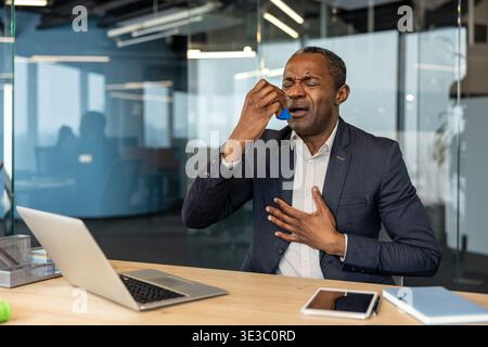 Mature businessman experiencing a respiratory attack, using an inhaler to alleviate breathing difficulties, placing a hand on his chest in an office setting Stock Photo