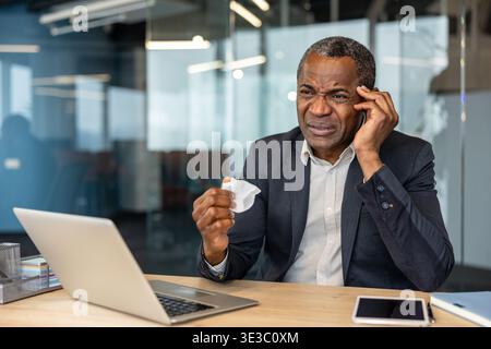 Mature professional man at his office desk rubbing his temple and holding a crumpled tissue, looking unwell and fatigued from headache, stress or flu while working on a laptop Stock Photo