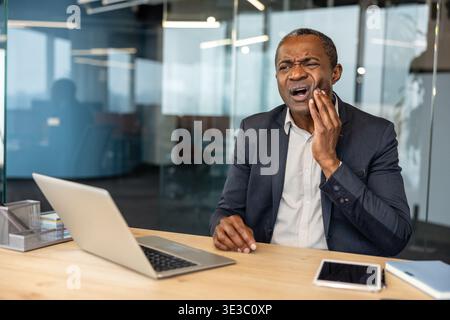 Mature man experiencing intense dental pain while working at his laptop in a contemporary office setting, highlighting discomfort interrupting productivity and health issues Stock Photo