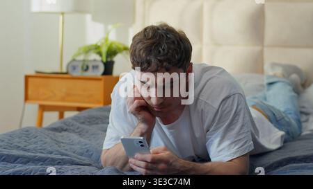 Worried Caucasian man using smartphone in bed at home frustrated male checking mobile phone problem error notification guy reading bad news banking Stock Photo
