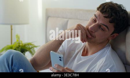 Tired upset Caucasian business man sitting bed in bedroom using mobile phone online stress work exhausted male guy overworked entrepreneur freelancer Stock Photo