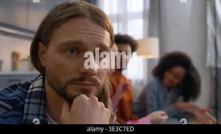 Despair Caucasian man at home worry thinking depressed male having panic attack feeling ignored while multiethnic African American friends talk Stock Photo