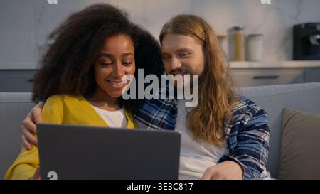 Multinational couple shopping online at home couch using laptop pc discussing purchase Caucasian man African American woman planning vacation Stock Photo