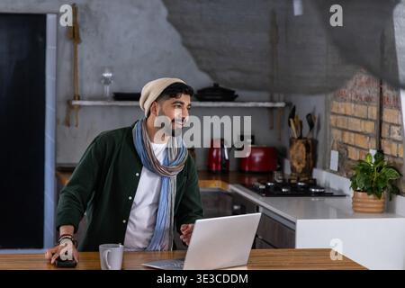 Bearded man early 30s wearing beanie scarf standing at kitchen island using laptop holding mug Stock Photo