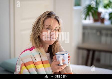 Cheerful tranquil woman with bed hair covered in blanket enjoys morning coffee sitting in comfy bed. Stock Photo