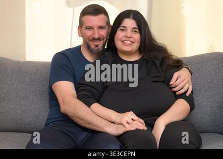 A man and woman sit on a couch together, smiling and embracing indoors. Stock Photo