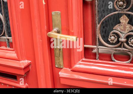 Close-up of vintage brass door handle on red wooden door with decorative metal grille. Stock Photo