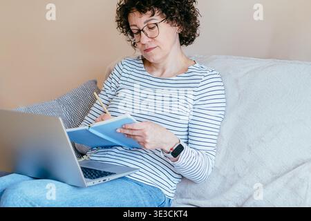 Focused middle aged woman planning in notebook with laptop at home Stock Photo