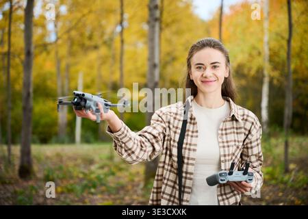 Woman launches drone from her hand while holding controller. Smiling young woman outdoors in autumn park, starting flight of quadcopter, concept of hobby, technology and modern lifestyle Stock Photo