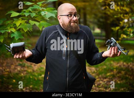 Man shrugs while holding drone and controller in hands. Bearded man looks confused in park comparing devices, unsure what to do, concept of choice, uncertainty and technology Stock Photo