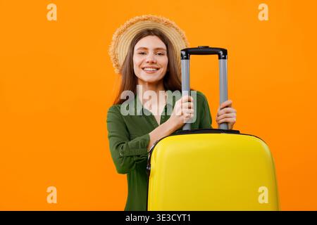 Woman With Yellow Suitcase and Straw Hat Stands Against Orange Background Ready for Travel Stock Photo