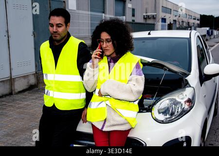 Man and woman wearing yellow high visibility vests standing by a broken down car with the hood open, woman making an emergency call on her smartphone Stock Photo