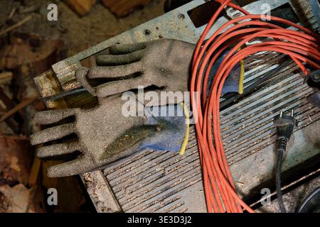 High-angle view of dirty, used work gloves with blue cuffs and a coiled red extension cord on a dusty metal workbench in a cluttered workshop Stock Photo