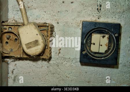 A dirty, extremely dusty vintage power outlet with a plugged-in old beige cord and a broken black light switch on a grimy, weathered workshop wall Stock Photo