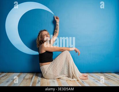 Young woman with dreadlocks smiling, sitting on floor reaching for a crescent moon projected on a blue wall Stock Photo