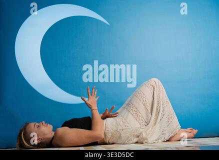 Woman lying down on floor in meditative pose with moon projected on blue background Stock Photo