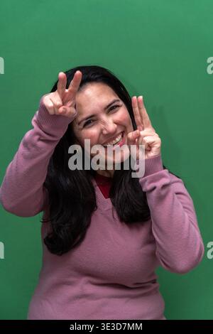 Woman standing on a green background, smiling brightly and showing peace signs with both hands, expressing joy and success Stock Photo