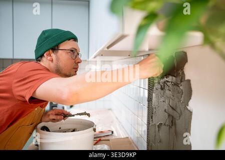 Man wearing a green beanie and glasses diligently installing new ceramic tiles on a kitchen wall, applying adhesive, and working on a diy home renovation project Stock Photo