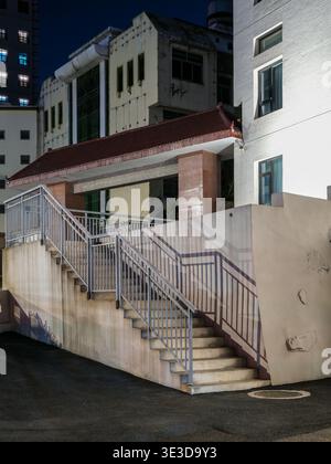 An outdoor concrete staircase leading to an old residential building with weathered walls illuminated by streetlights in a dark alley at night. The sc Stock Photo