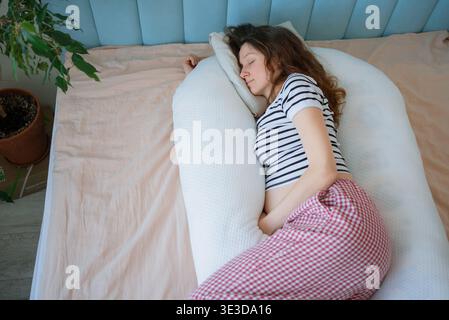 Pregnant woman sleeping deeply on her side in bed, supported by a large u-shaped maternity pillow, ensuring optimal comfort and body alignment for restful nights during pregnancy Stock Photo