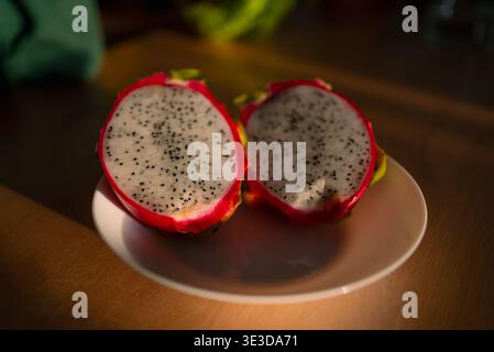 Halved dragon fruit revealing its distinctive white flesh speckled with tiny black seeds and bright pink skin, resting on a white plate, symbolizing fresh, healthy, and exotic fruit Stock Photo