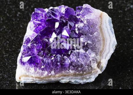 Sample of a raw geode made of amethyst mineral crystals on a black granite background, close-up from Russia Stock Photo